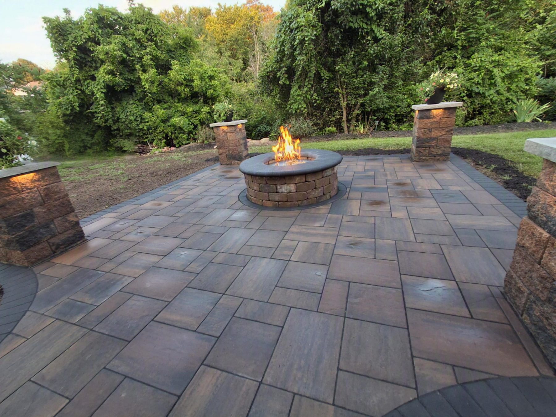 A gas fire pit burning on a paver patio between lit stone pillars, built by M.S.S Landscaping